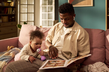 Black middle aged man sitting on sofa reading picture book to toddler girl while spending time together during paternity leave, toddler girl holding purple toy and looking at book