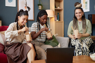 Three young adult women, including Black and Caucasian, sitting on sofa eating noodles with chopsticks while watching laptop screen, smiling and engaging in casual conversation together
