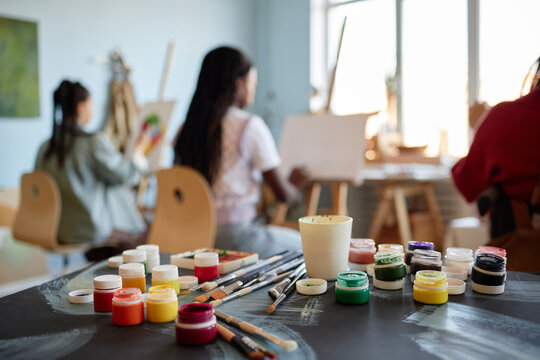 Multiethnic group of young women sitting at easels painting on canvases in art studio, various paint jars and brushes arranged on table in foreground, focused on creative activity - Powered by Adobe