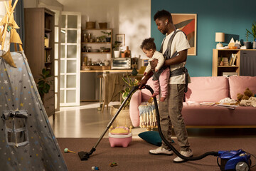 Black young adult man vacuuming living room floor while carrying toddler child in baby carrier,...