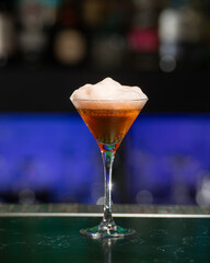 A deep amber or brown alcoholic cocktail, topped with a thick layer of white foam, is presented in...