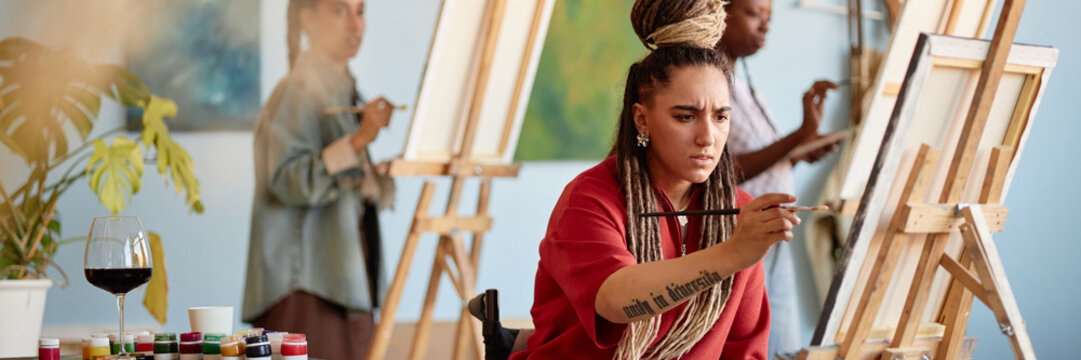 Header of young adult Caucasian woman in wheelchair painting on canvas in art studio, holding paintbrush with focused expression, two other women painting on easels in background - Powered by Adobe