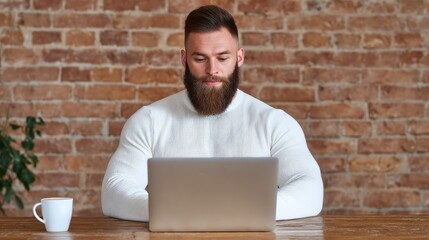 A bearded man in a white sweater works on a laptop at a wooden table with a coffee cup nearby, set against a brick wall background.