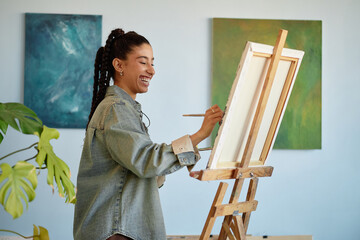 Young adult woman smiling while painting on canvas at easel, holding paintbrush in hand, standing...