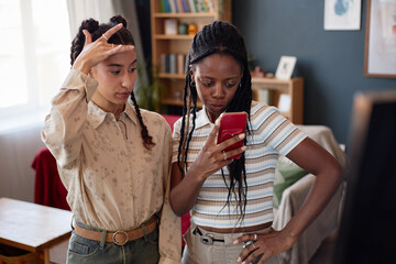 Two young adult multiethnic women standing together looking at smartphone screen, appearing focused and engaged in discussion in modern home setting