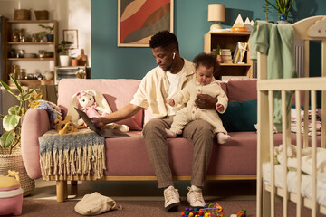 Black young adult man sitting on sofa holding infant child, while working on laptop multitasking during paternity leave in home setting with toys and crib visible in background
