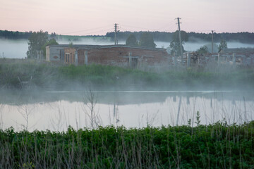 Misty landscape featuring abandoned buildings near a tranquil water body, surrounded by lush...