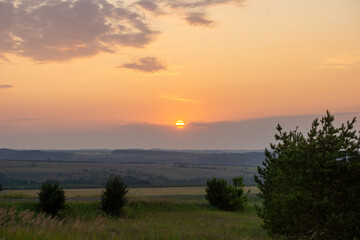 Sunset over rolling hills with vibrant orange and yellow hues illuminating the sky, framed by lush green trees and tall grass, creating a serene natural landscape
