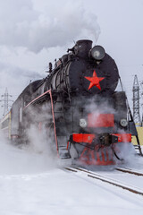 Vintage steam locomotive with red star emblem is releasing steam while traveling on snowy tracks, surrounded by power lines and a winter landscape, showcasing historical transportation