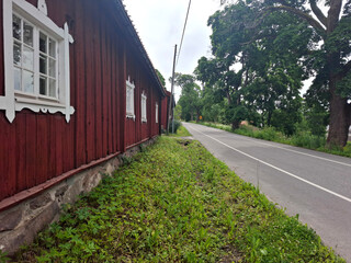 Obraz premium Country road beside a traditional nordic red wooden house with white window frames. Lush green trees line the quiet rural street under a cloudy summer sky.