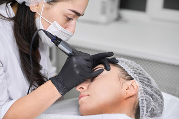 Skilled technician applies permanent eyebrow makeup to young woman in calm salon. With black...