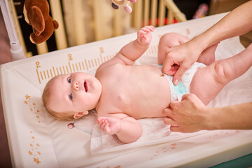 Infant with light hair lies on changing table. Adult mother hands adjust white nappy. Soft lighting...