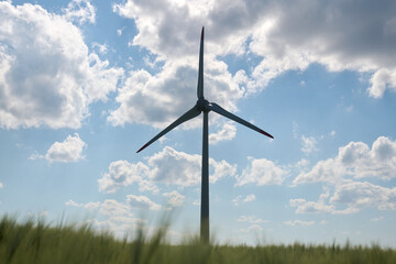 Single wind turbine silhouetted against bright blue sky with scattered fluffy clouds and blurred...