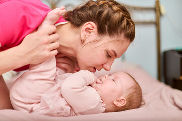 Young woman with brown hair kisses infant daughter with light hair on bed. Warm, gentle moment...
