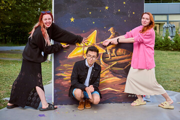Two young female artists and young male pose playfully with outdoor painting in park. Vibrant...