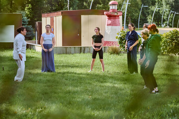 Group of adults practicing qigong in grassy garden. Female teacher leading session under sunny sky....