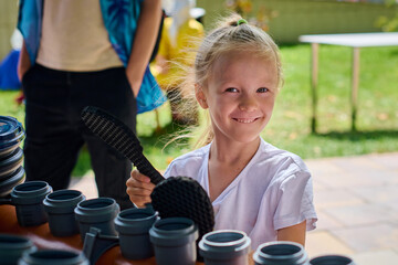 Smiling young girl with blonde hair playing PVC pipe instrument outdoors. Sunny day, colorful...