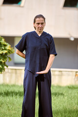 Mature caucasian woman with brown hair engages in qigong in sunlit park. Wearing dark attire, standing on vibrant grass, focused expression in peaceful outdoor setting