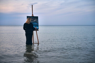 Young woman stands in serene ocean waters at dusk, beside easel displaying painting. Scene captures blend of creativity and nature, evoking sense of calm and inspiration