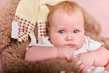 Baby girl, light hair, caucasian, lying on pink blanket, cuddling toy bear. Soft lighting adds warmth, creating cozy and serene atmosphere, with focused gaze