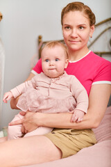 Young mother with blonde hair cradling baby daughter in cozy room. Soft lighting creates warm atmosphere. Mother wears pink top while daughter is in pink dress, both looking content
