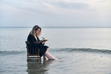 Young woman sits on chair in shallow water at sunset, writing in notebook. Serene ocean setting evokes feelings of peace and introspection, blending creativity and nature