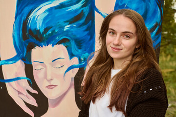 Young woman with long brown hair and fair skin stands beside art piece. Blue dominates painting. Outdoor setting, natural lighting enhances vivid hues