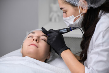 Young woman receives eyebrow permanent makeup in serene spa. Beauty technician, wearing gloves, focuses intently, creating calm, professional atmosphere