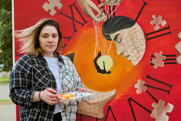 Young woman holding paint palette stands by vibrant surreal clock painting with puzzle patterns. Outdoor scene filled with creativity and orange hues
