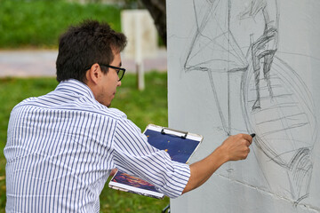 Young asian male artist sketching with pencil on outdoor wall. Focused expression, striped shirt, glasses. Background of lush grass and trees. Side view