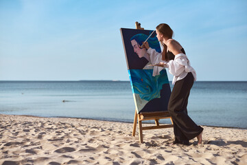 Young woman artist paints vibrant portrait on sandy beach, capturing tranquil ambiance of seaside. Her focused expression and calm sea evoke creativity and peace