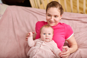 Young adult female with brown hair holding adorable baby girl in pink outfit. European ethnicity, both smiling indoors on soft pink blanket with wooden crib nearby