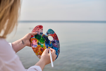 Young female artist blends vibrant paint on palette by serene seaside. Calming ambiance and natural light inspire creativity and introspection
