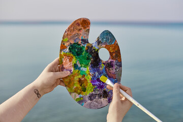 Young female holds colorful artist palette against tranquil seascape, symbolizing creativity and inspiration drawn from nature beauty. Blend of hues evokes peace