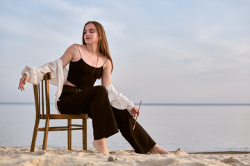 Young female painter sits on wooden chair on sandy beach, holding paintbrush. Serene ocean background enhances peaceful mood, evoking calm inspiration