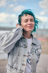 Young white woman with short blue hair in denim jacket outdoors. Soft smile, relaxed pose, cloudy sky background, windy day, natural lighting