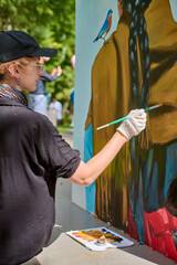 Young adult female artist with gloves and cap painting vibrant mural. Focused, intent expression. Sunlit park backdrop with greenery enhances creativity