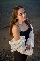 Young woman stands serenely by shoreline, draped in white shirt over black outfit. Setting sun...