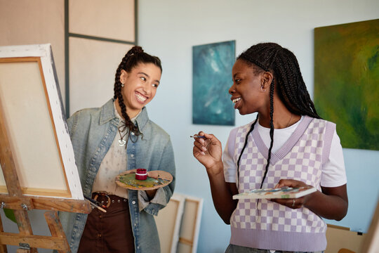 Diverse women painting together in art studio, smiling and interacting while holding paintbrushes and palettes, standing near easel - Powered by Adobe