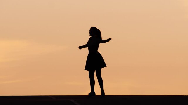On hilltop against sunset sky, young woman's silhouette dances with joyful abandon. Her shadow figure spins and twirls, arms outstretched, her dress skirt fly around.