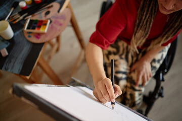 Young Caucasian woman with disability sitting in wheelchair drawing on canvas using pencil, holding sketchbook with left hand, art supplies and palette visible on table nearby