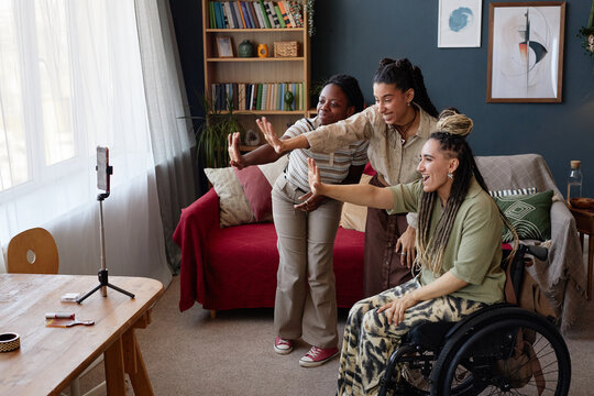 Diverse group of young adult women smiling and waving at smartphone camera, Black woman, multiethnic woman, and Caucasian woman with disability in wheelchair recording video together