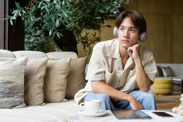 Non binary Asian student man sitting in modern coffee shop wearing headphones