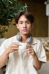 Non binary Asian student man sitting in modern cafe drinking coffee