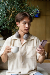 Non binary Asian student man sitting in modern cafe using phone and drinking coffee
