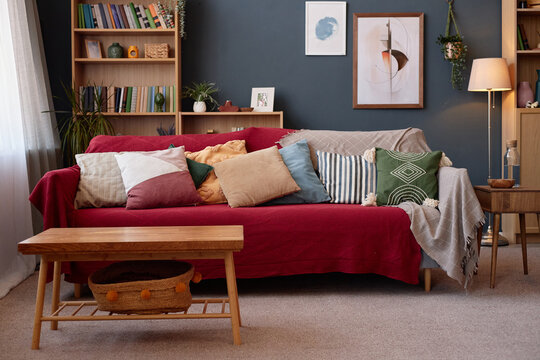 Empty living room scene showing wooden coffee table in foreground, red sofa with assorted pillows and bookshelves in background, no people visible, cozy home interior