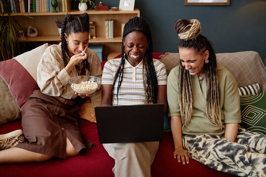 Three young adult women, including Black and Caucasian, sitting on sofa laughing and watching laptop screen together, one holding bowl of popcorn, enjoying shared entertainment - Powered by Adobe