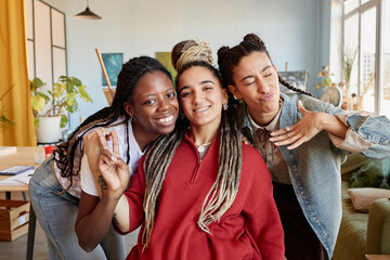 Three young adult women, including Black and Caucasian ethnicities, smiling and posing together indoors, one woman making peace sign while another gestures playfully toward camera