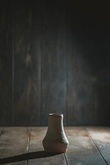 Handcrafted ceramic vase rests on a rustic wooden surface, illuminated by soft, dramatic lighting. The dark background enhances the focus on the vase's unique shape and texture