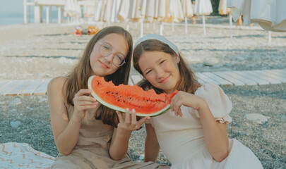 Two smiling girls sitting on the beach holding a juicy watermelon slice, enjoying a sunny summer...